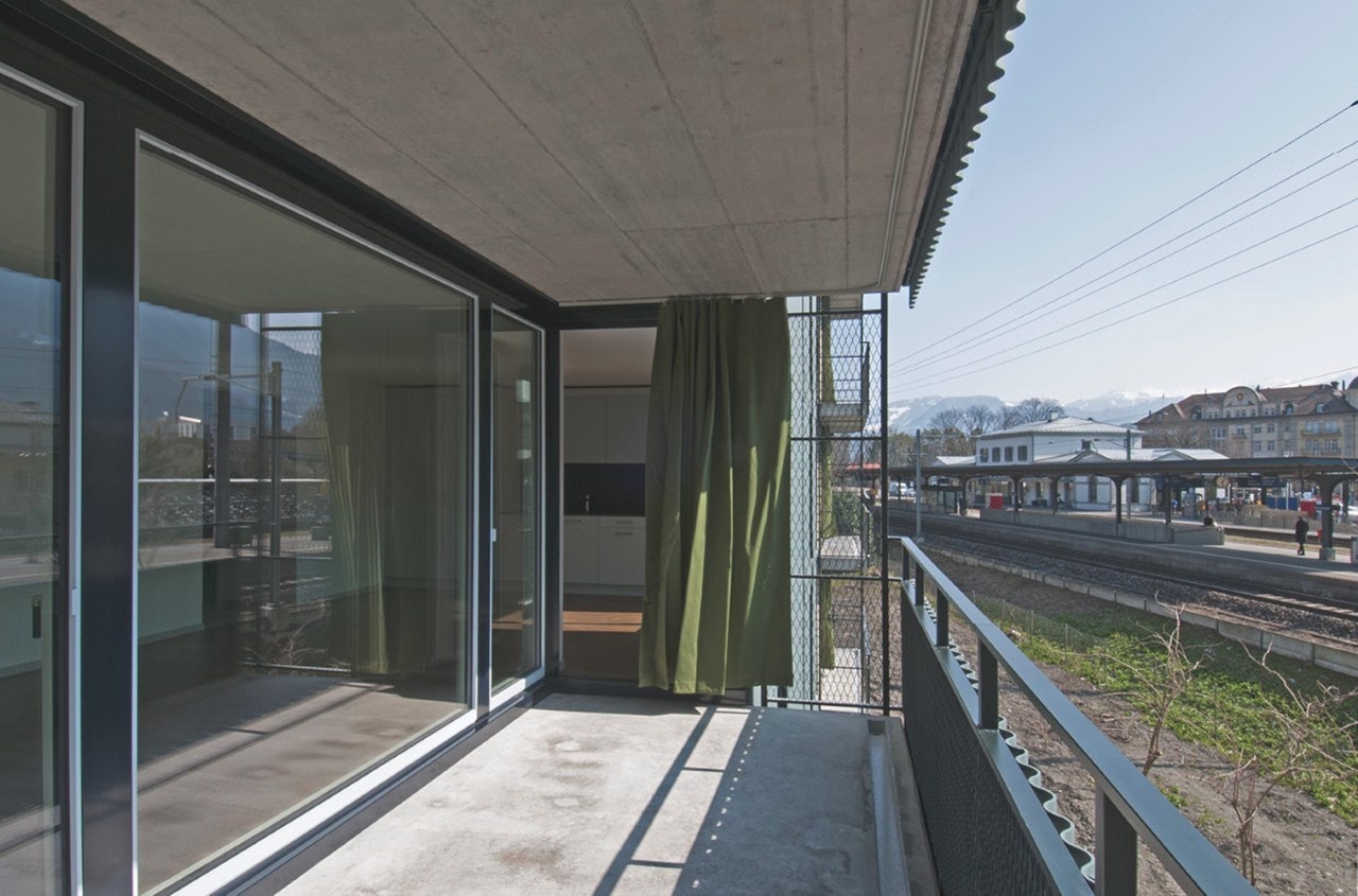 Concrete balcony with metal and chain link railings and a green curtain. Floor to ceiling windows form the back wall of the balcony.