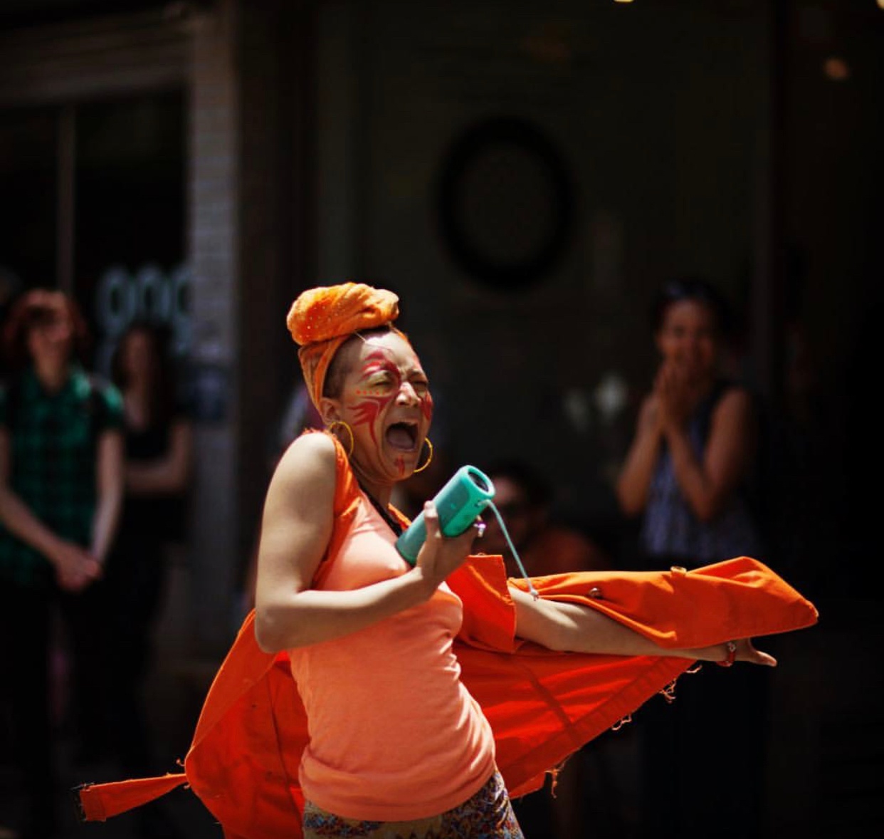A black and Native nonbinary femme person performing for an audience that stands in the background. They wear an orange tank top and orange headscarf and an orange sweatshirt spins out from their body like a cape. 