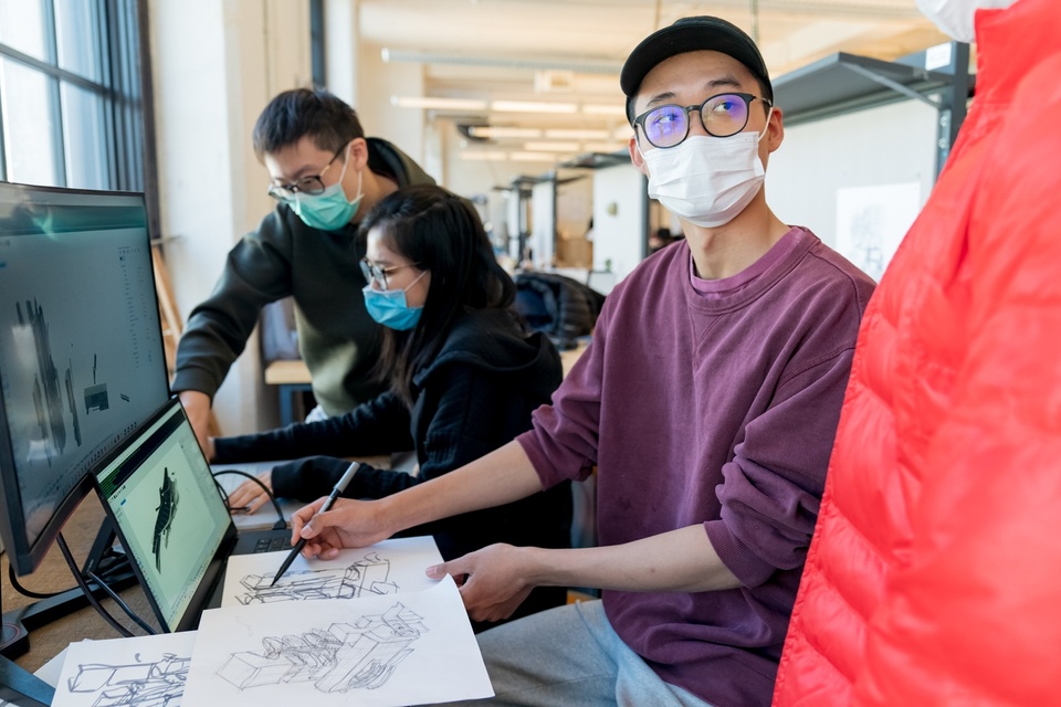 Person in a ballcap looks up from an architectural sketch they are making of a boxy machine in front of a laptop displaying a reference image.