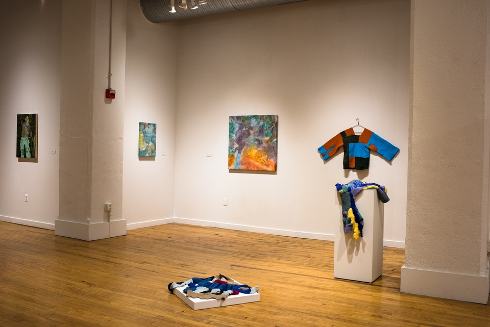 View of a gallery space with several paintings and three knitted items on display.