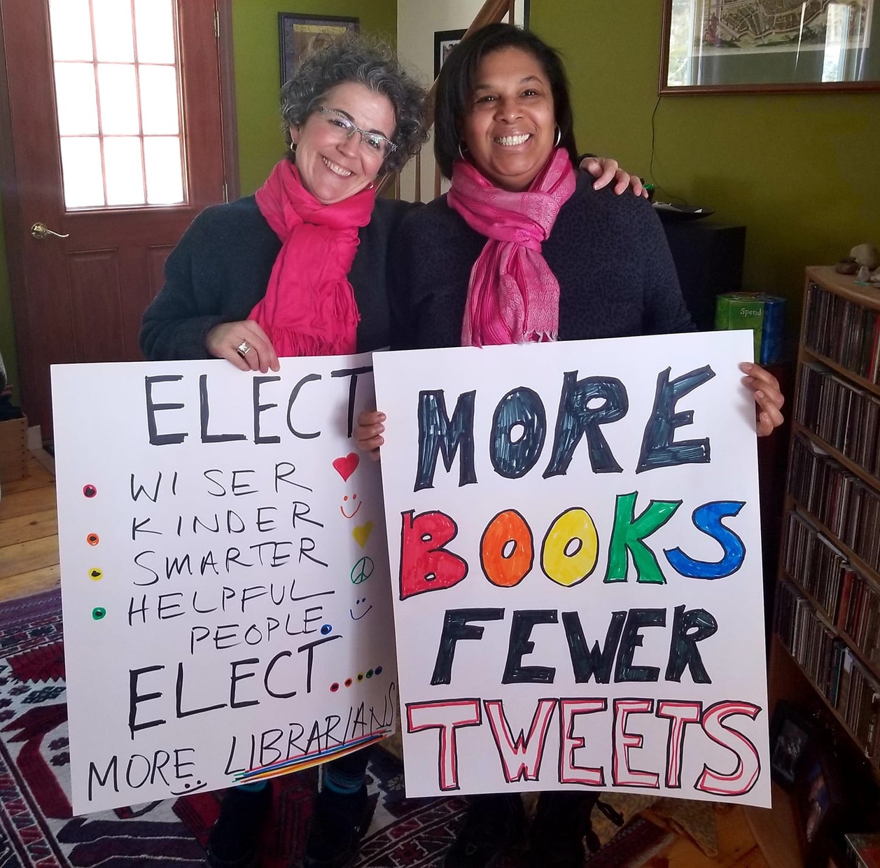 Two middle-aged women standing together in an interior and wearing pink scarves are smiling and holding posters that say “ELECT… MORE LIBRARIANS” and “MORE BOOKS FEWER TWEETS.”