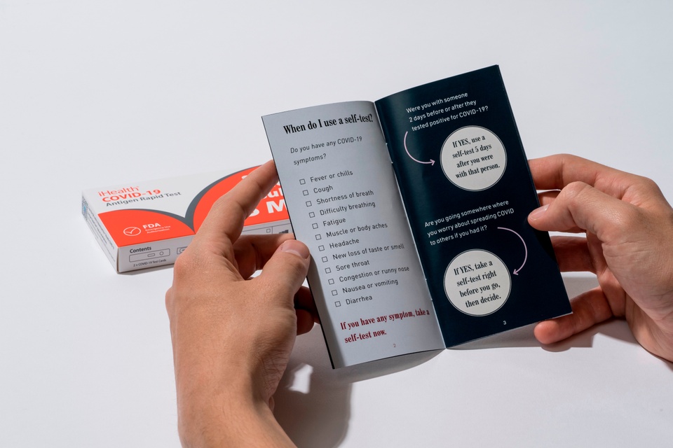 An iHealth self-test kit lies open behind the user, who has opened an instructional booklet. The left page reads a list of symptoms, while the right page asks two questions about contact with someone diagnosed with COVID.