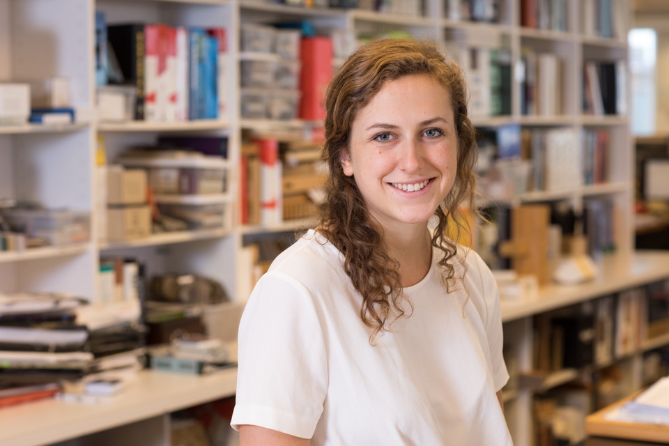 A person wearing a white shirt looks at the camera and smiles. In the background are bookshelves.