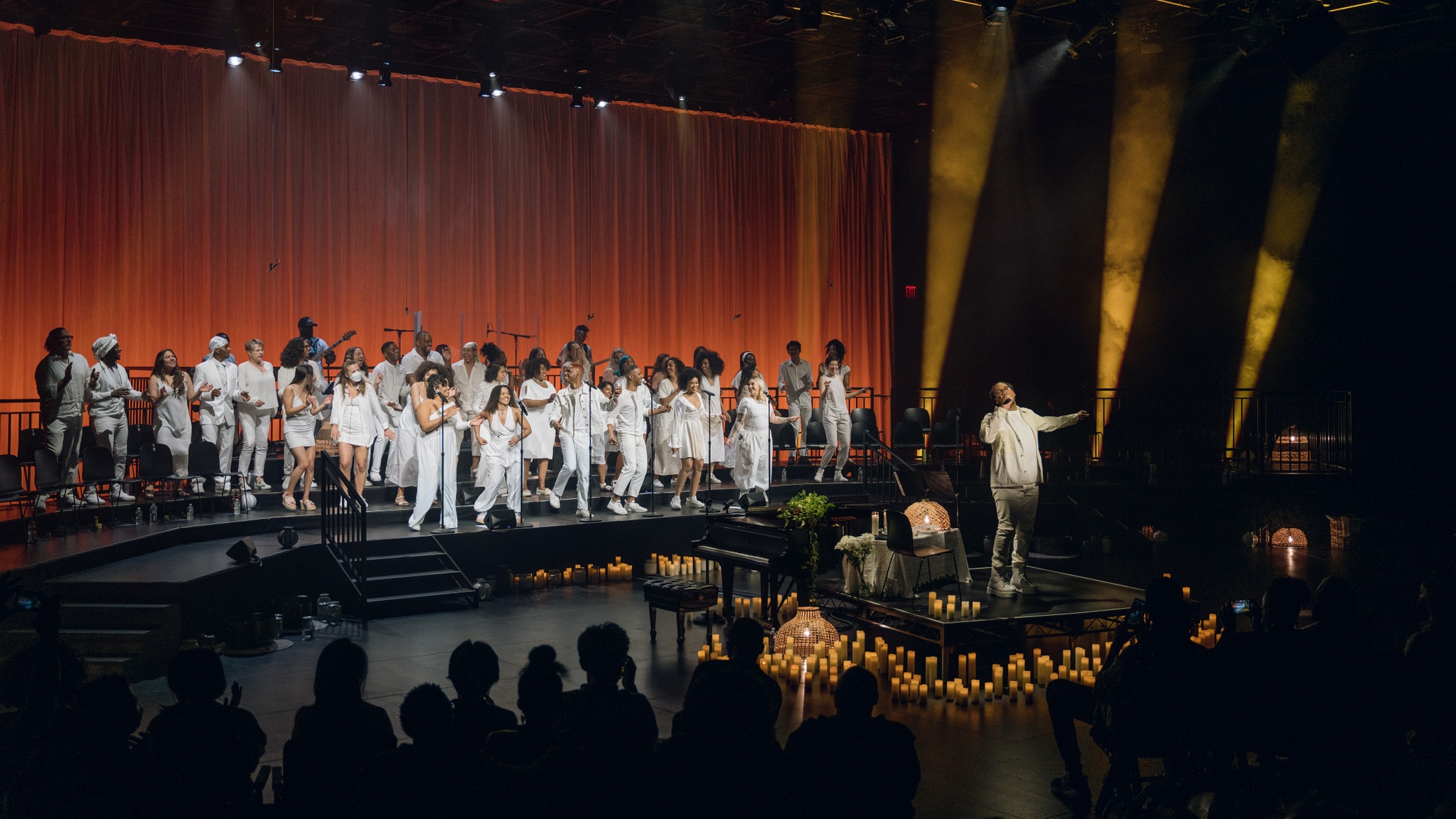 Troy Anthony, a Black man dressed in white, stands on a small stage in a black box theater with a choir on risers behind him. Troy stretches out his hands and the choir members dance. They are also dressed all in white. Around Troy are numerous lit candles. Behind the choir, a curtain is lit in deep umber tones.