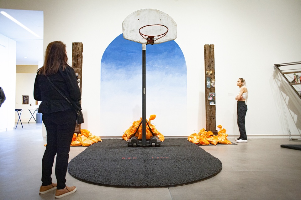Installation of a basketball hoop missing its net, set into a small section of rubber play area. On the wall behind it is an ombre painting of sky in an arched shape. Flanking it are two upright railroad ties with photographs nailed to them.