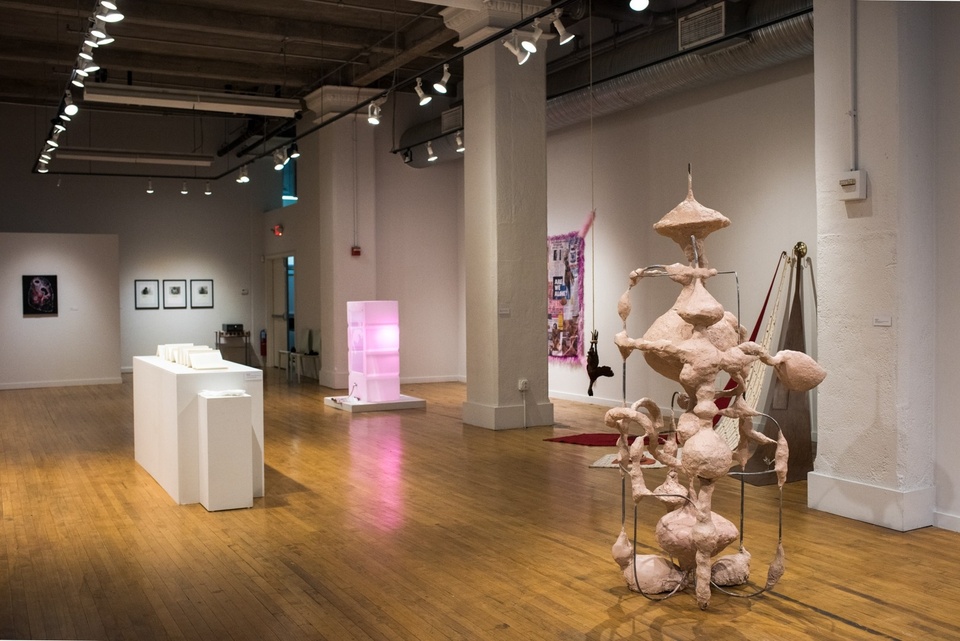View of gallery featuring sculptural and 2D work. In the foreground is a peach-color head-height plaster sculpture. In the background can be seen a pink glowing box.