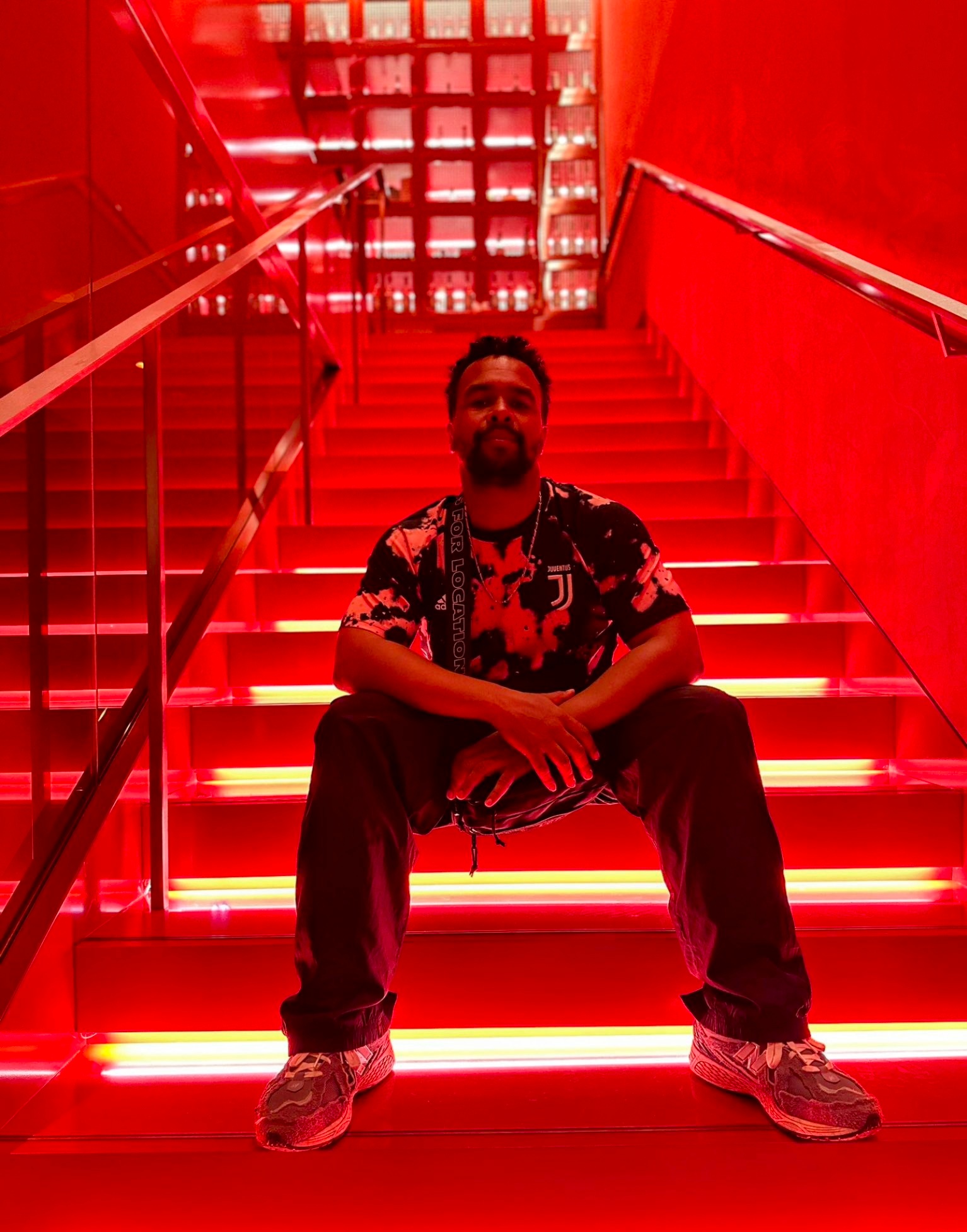 A Black man sits on a staircase suffused with red atmospheric light. He looks at us from slightly above on the stairs where he sits with hands crossed over his legs. He wears a floral print shirt, pants, and sneakers.