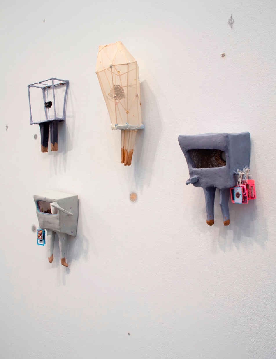 A group of small, abstract, muted-colored sculptures, with a few spots of bright color, hang on a white wall covered with small grey speckles.