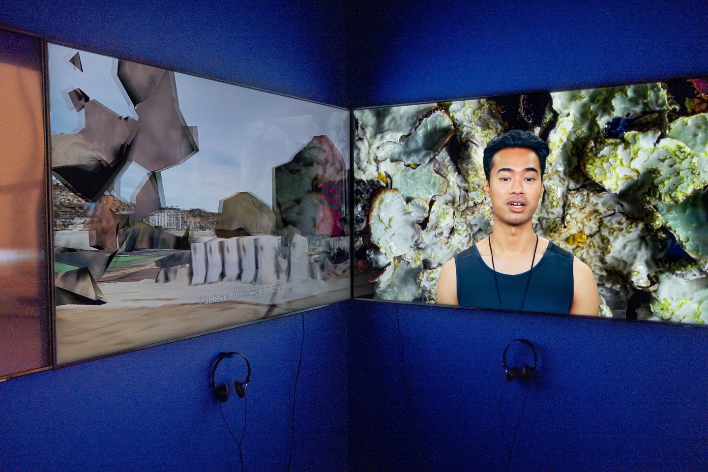 Two large screen monitors mounted on a blue wall with headphones hanging underneath. Images of a building and a person talking are on the screens.