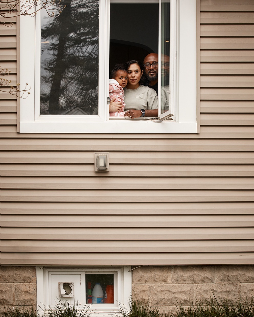 A photograph of a dark-skinned family, one adult male, one adult female, and one child, look out of a white framed window on a tan house.