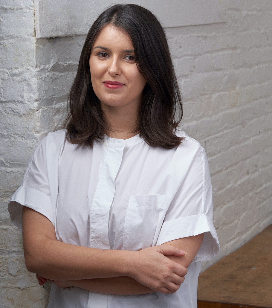 A portrait of Anna Harsanyi who poses against a white brick wall. She wears a white short sleeve dress with her arms crossed across her waist. She has dark brown shoulder length hair and and looks at us while smiling. 