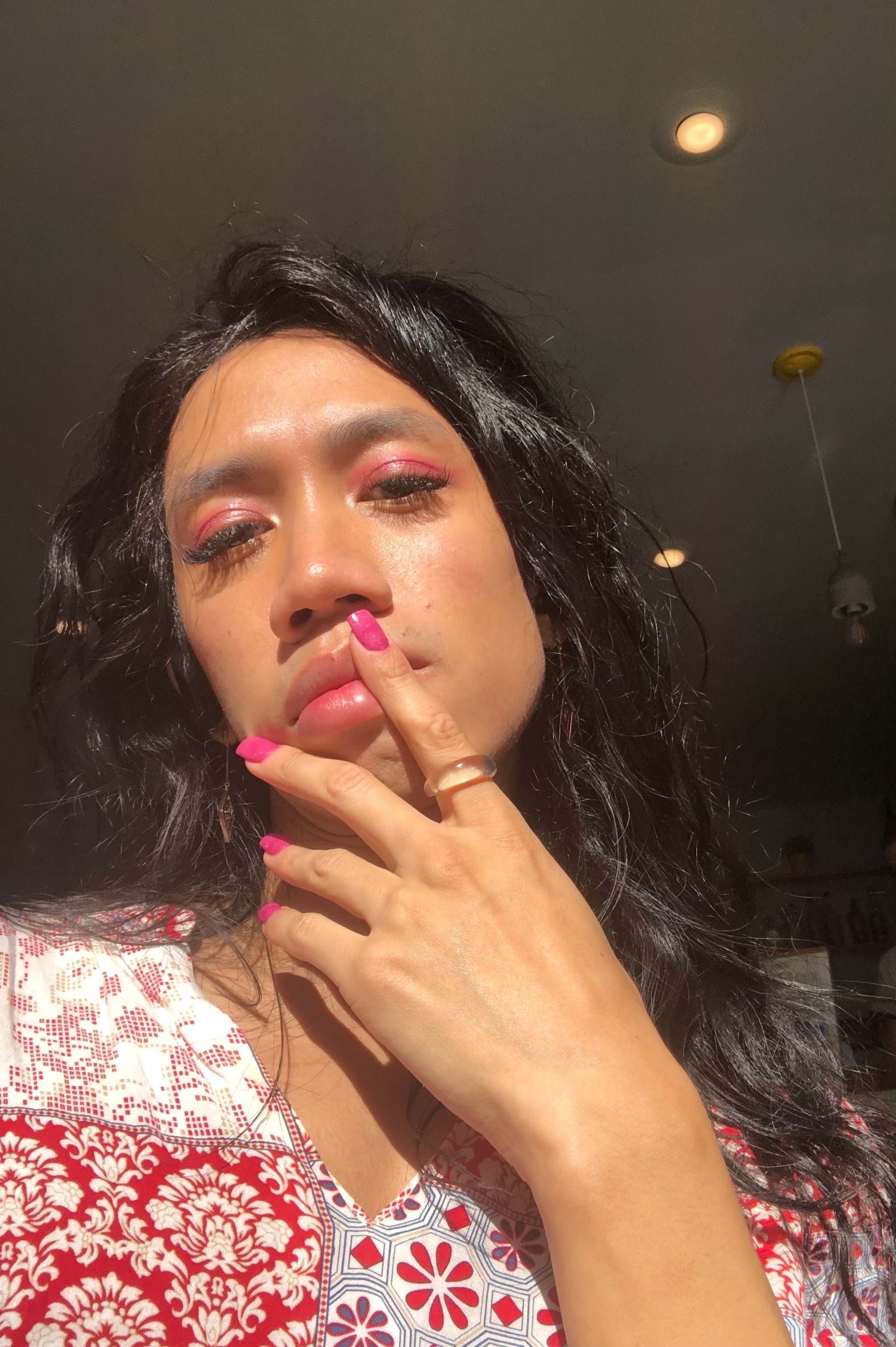A portrait of Benedict Nguyễn who is seen from an angle below as if in a selfie. Benedict holds two fingers to their mouth, while wearing matching pink nail polish and lipstick. They have long dark hair that falls beneath their shoulders. 