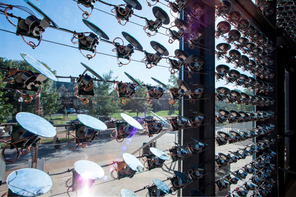 Window with many rows of small, round mirrors strung on wires and connected to individual motors with a rotating joint, controlling the angle of reflection.