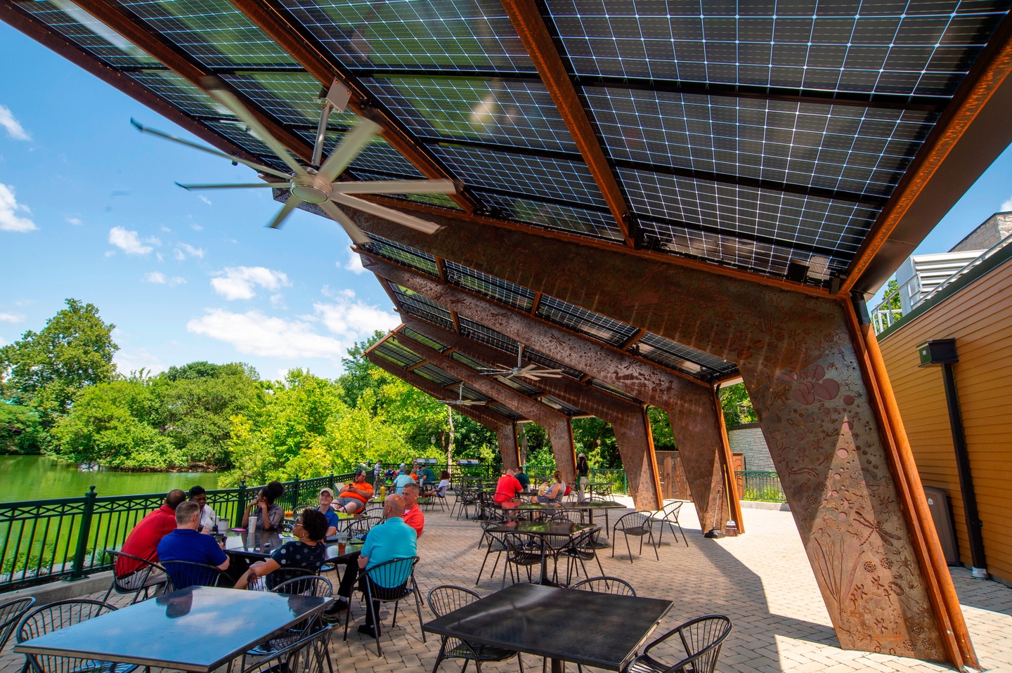 Outdoor photo of people seated on a patio at small, square tables under a solar shade canopy, made of angled supports with rich wood and solar panels above. A body of water can be seen in the distance.