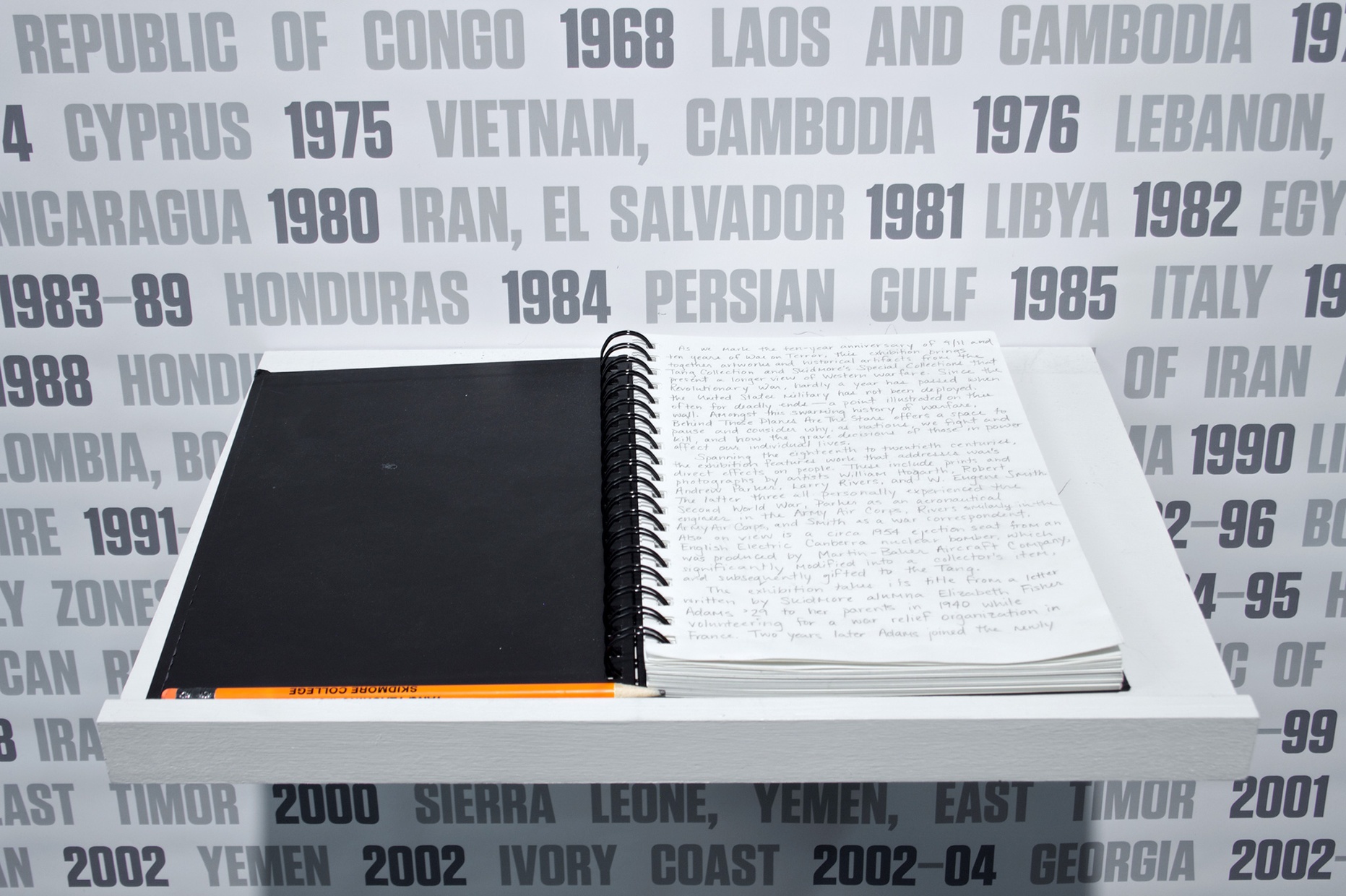 An open notebook sits on a white shelf with pencil notes. The wall behind lists names of countries with date-ranges next to the country.