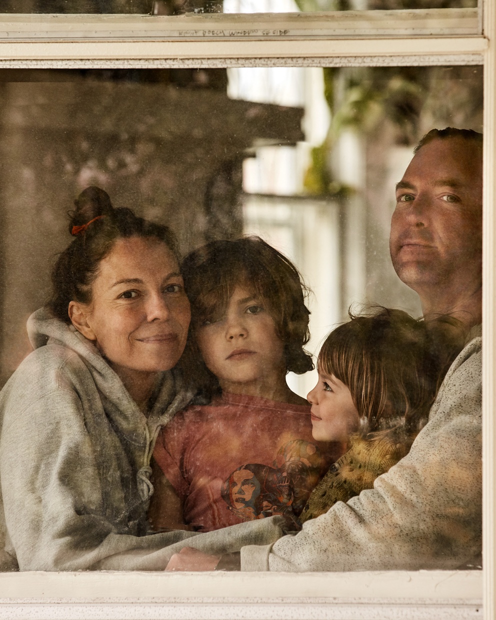 A photograph of a light-skinned family, one adult male and adult female, and two children, look out from a white-framed window.