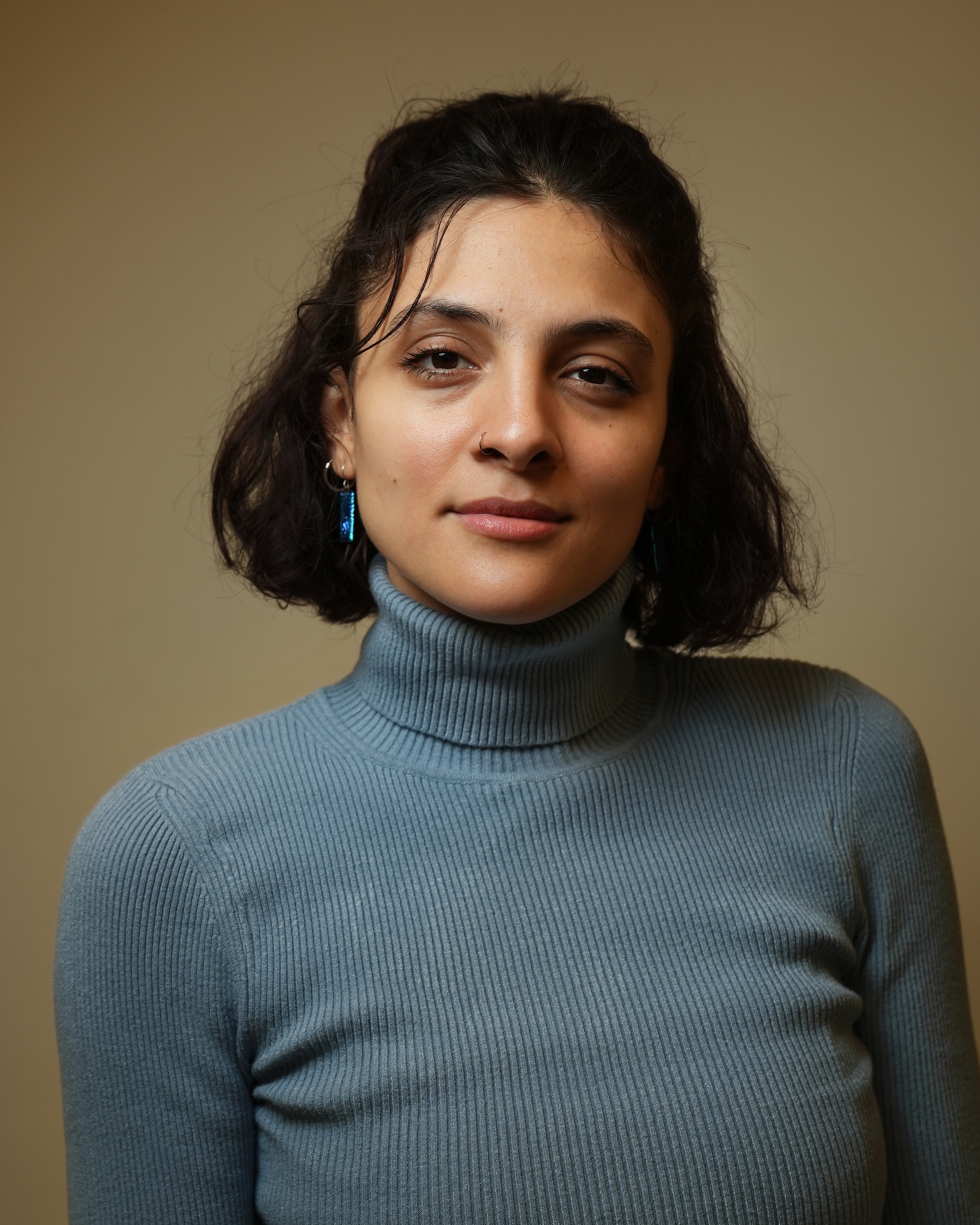 A portrait of Aya Aziz against a neutral brown background. Aya has short dark brown hair that is chin length and pulled half back. She wears a light blue tutrtleneck and looks directly at us, with a slight smile forming on her face. 