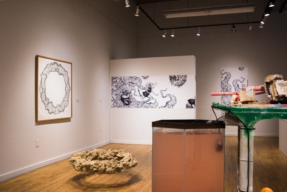 Small gallery space; in the foreground is a tank of murky orange liquid hooked up to a construction of tubes; in the background are two black and white drawings and on the floor is a sand-colored donut shaped sculpture.