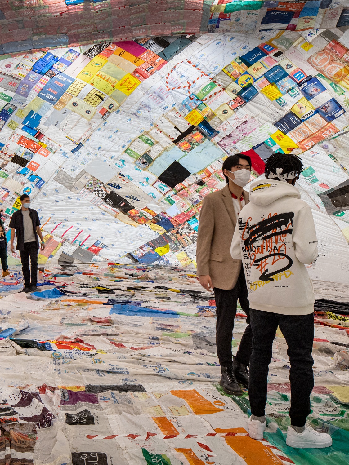 Three people stand inside a cavernous ballon made of a colorful patchwork of reused plastic bags quilted together with tape