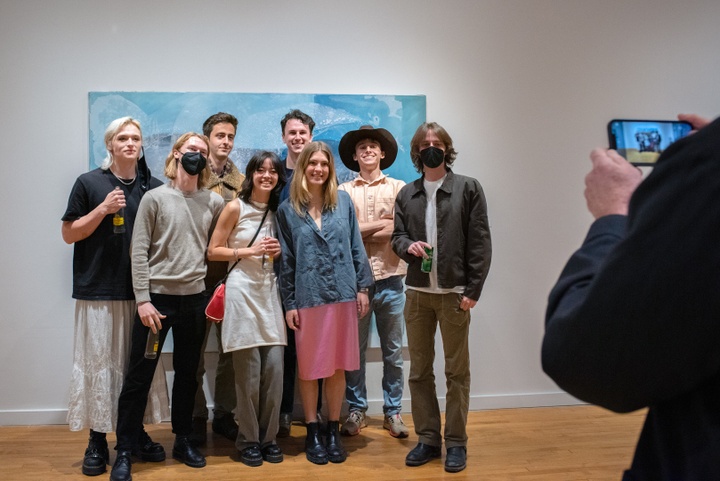 Group of smiling people pose in front of a teal painting in a gallery while someone takes their photo.