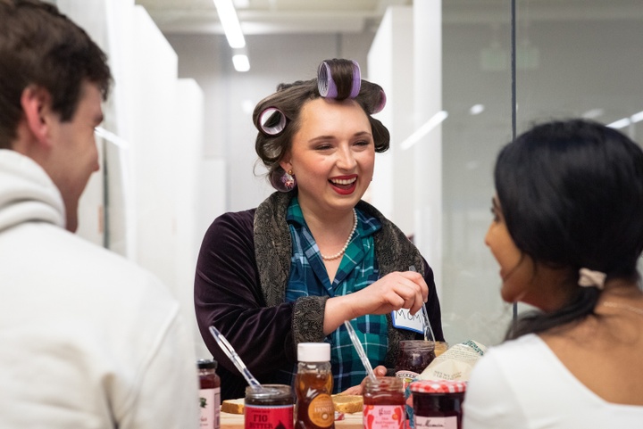 Person in hair rollers and bright red lipstick smiles sweetly at people across the table, while opening a jar of jam.