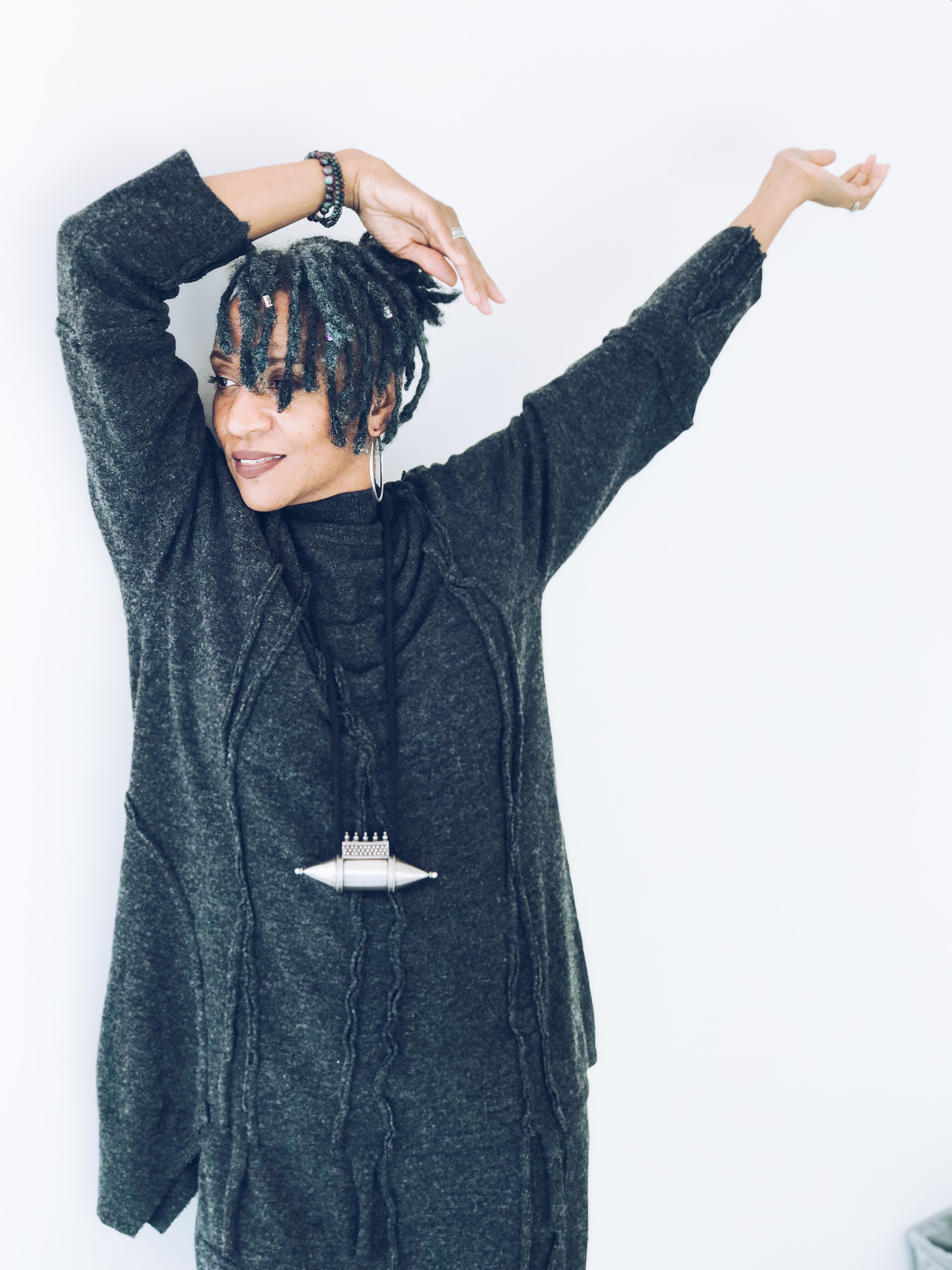 A Black woman with short locs falling over her eyes raises her hands above her head as if in joyful relaxation. She wears a long gray sweater and poses in front of a white wall.