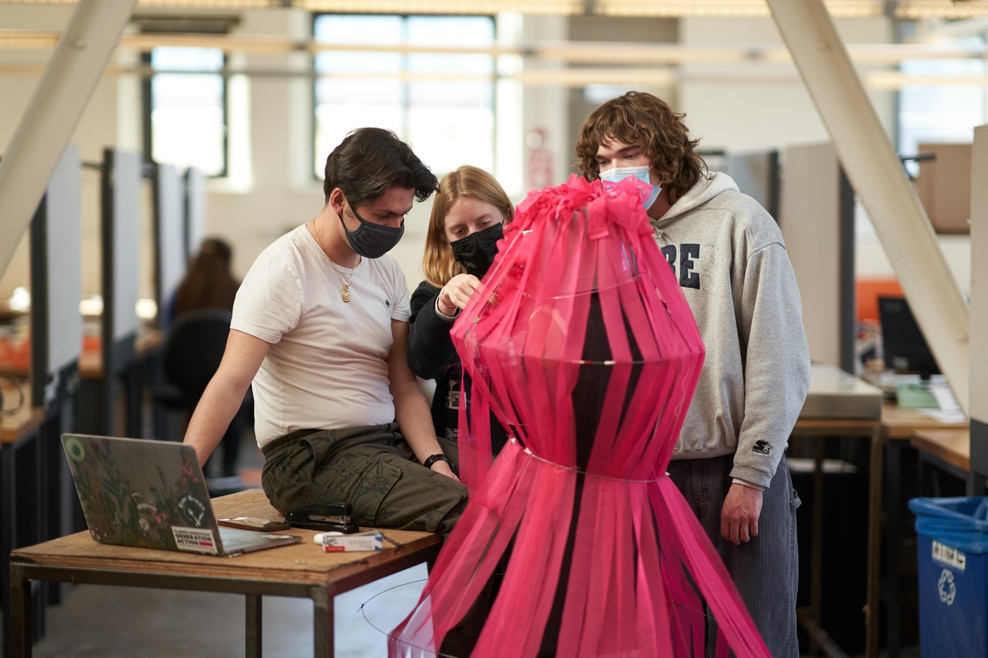 Two students stand around another one, helping arrange a wire-like dome structure around the middle student's head.