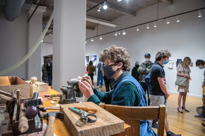 Person sits at a carpenter's bench in a gallery space and carves a small white block of soap into an animal while people mill around behind them.