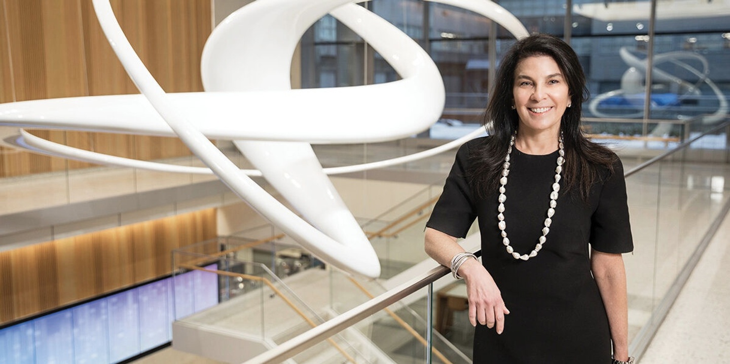 Photo of Vicki Match Suna, standing against a metal railing with clear glass next to a staircase. In the background is a large, swirly white sculpture, which is reflected in the windows behind her.