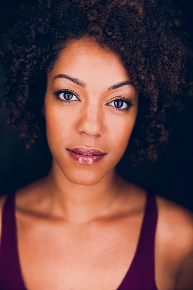 A portrait of actor Nemuna Ceesay. She has light brown skin, curly brown hair, wears a tank top, and arches one eyebrow at the camera.