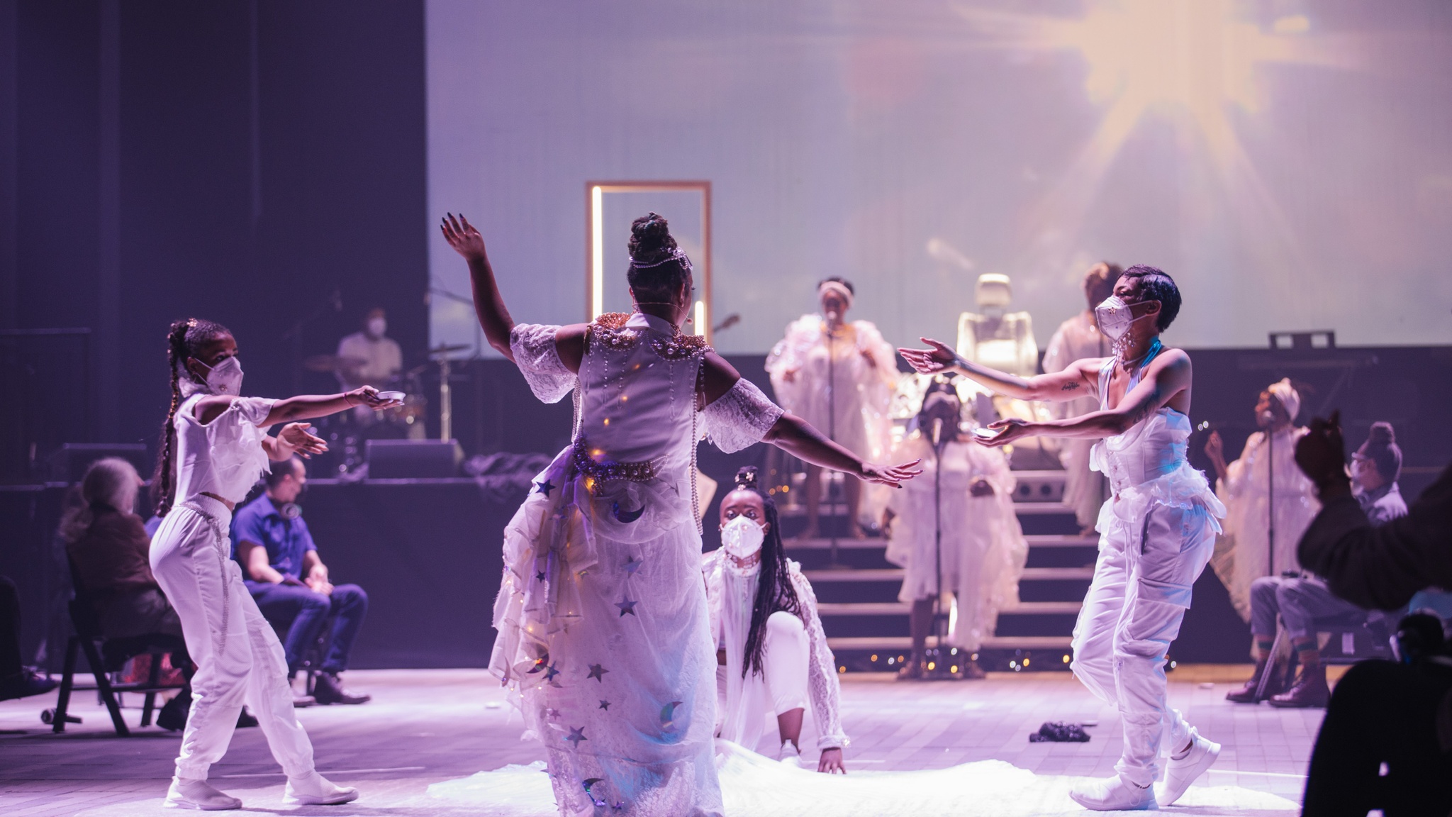 Four performers dressed all in white stand in a circle at the center of a performance space. In the background, additional performers in white stand on stairs leading up to a stage. The set and costume design include small twinkling lights evoking a cosmic scene. 
