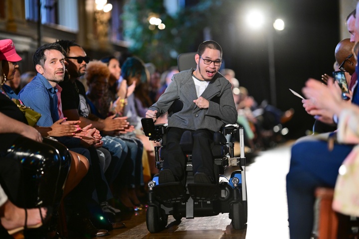 A model moves down the runway in a wheelchair wearing a grey jacket