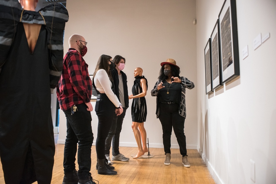 Person presents to a row of listeners in front of a mannequin in a black ribbon dress and three framed fabric pieces on a gallery wall.