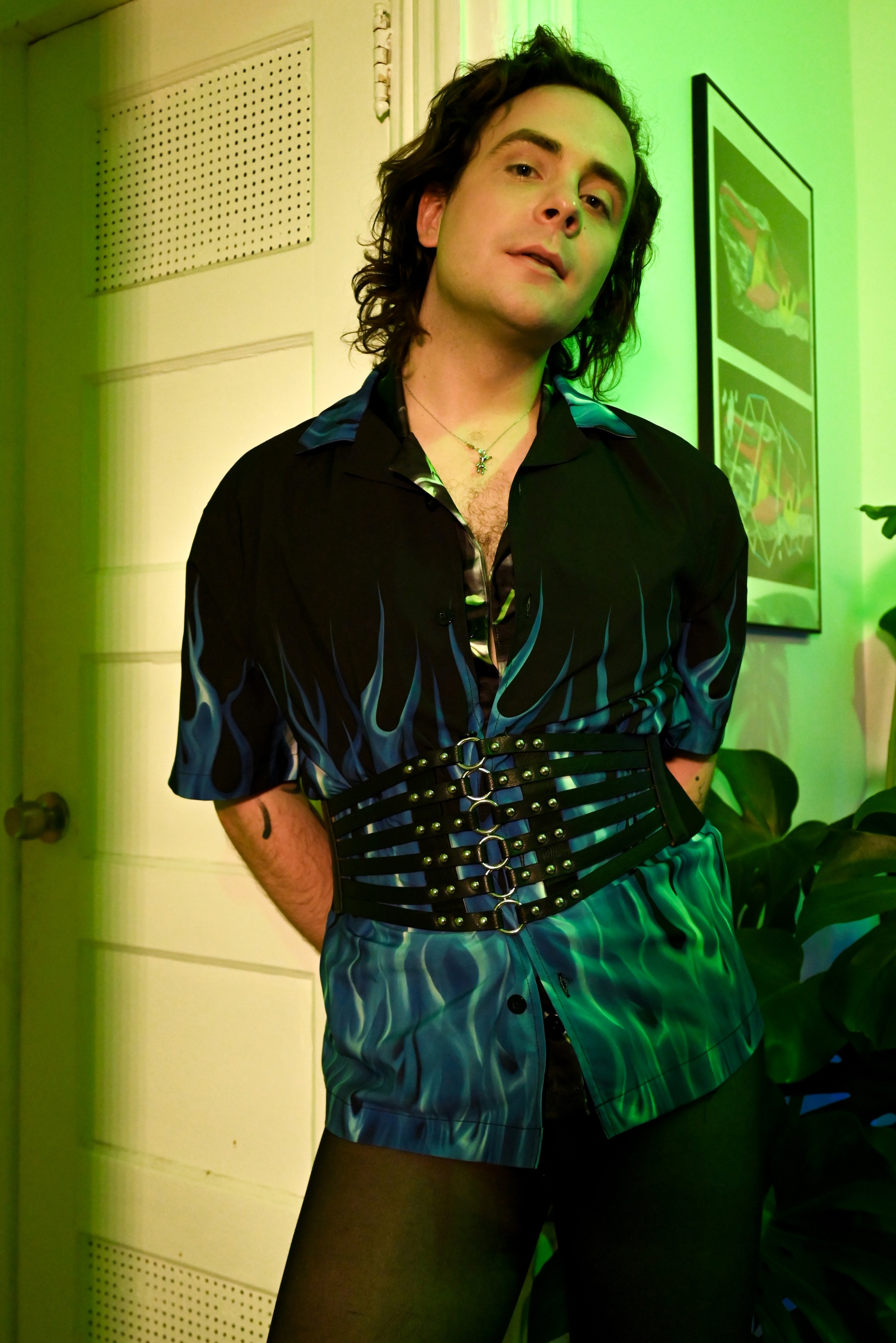 Electronic musician Despina stands in a doorway with green light in the room behind them. They hold their head slightly cocked backward and wear a short sleeve shirt with a print of blue flames rising from the bottom hem. Around their waste is a wide leather belt with metal rings down the center. 