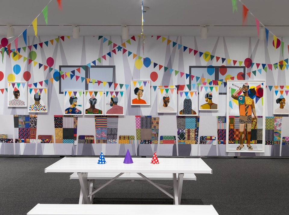 A multi-part installation with a full-wall mural portrays a joyful party scene. The installation comprises framed portraits of Black individuals in vibrant, patterned garments; balloons and colorful bunting (real and painted); and, in the foreground, a white picnic table with blue, orange, and purple party hats. The paintings are executed in a graphic style, with segmented of color blocks reminiscent of stained glass.  