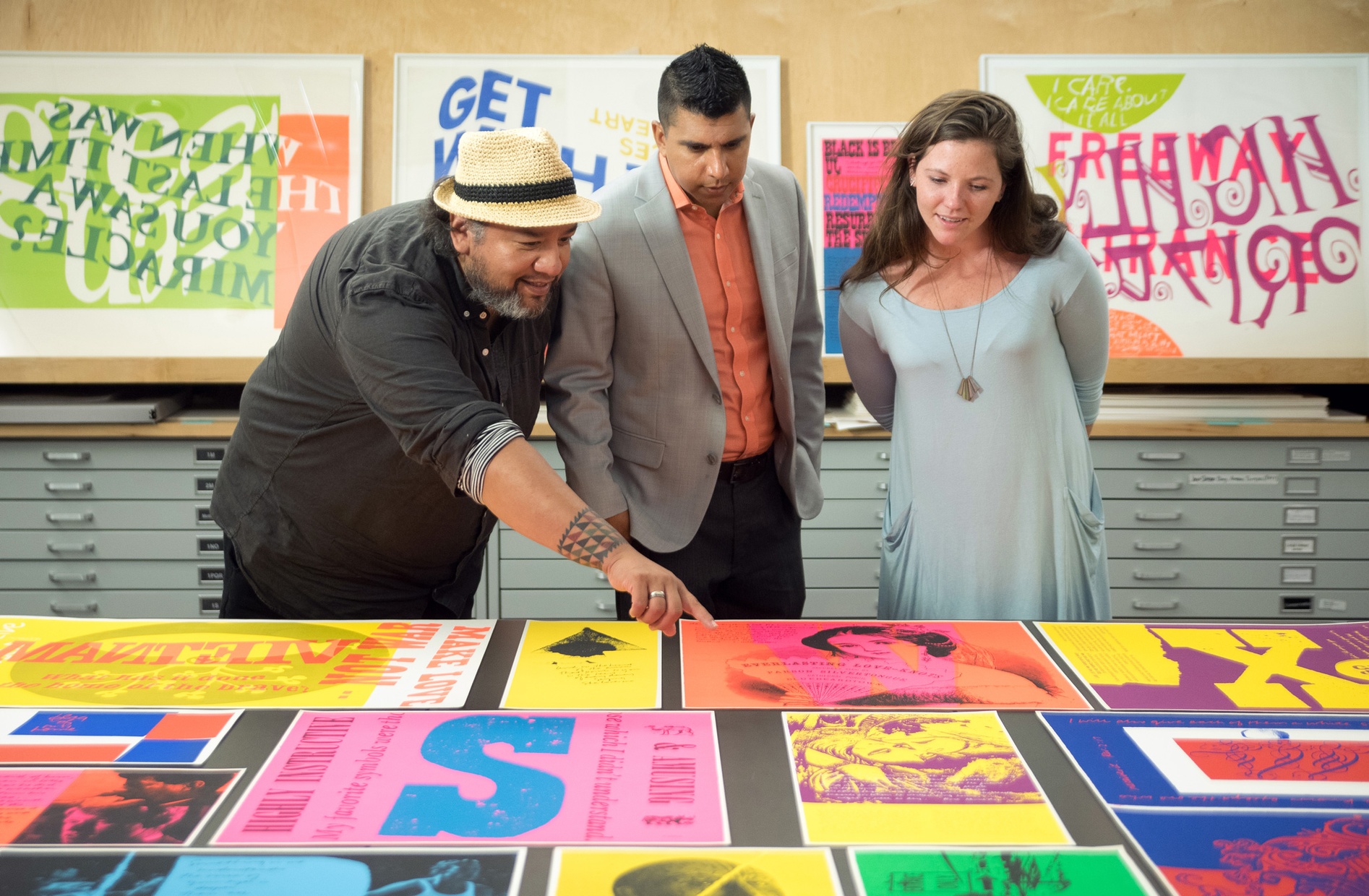 Three people look down at a table covered in colorful screenprints. One of them, a tanned, middle-aged, Native man points to the prints.
