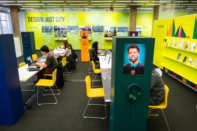People working at two separate long, white tables in a room with light orange chairs and neon yellow walls. In front of each table is a bold-colored rectangular station; the one on the right is forest green and includes headphones, as well as a video monitor with a person on screen.
