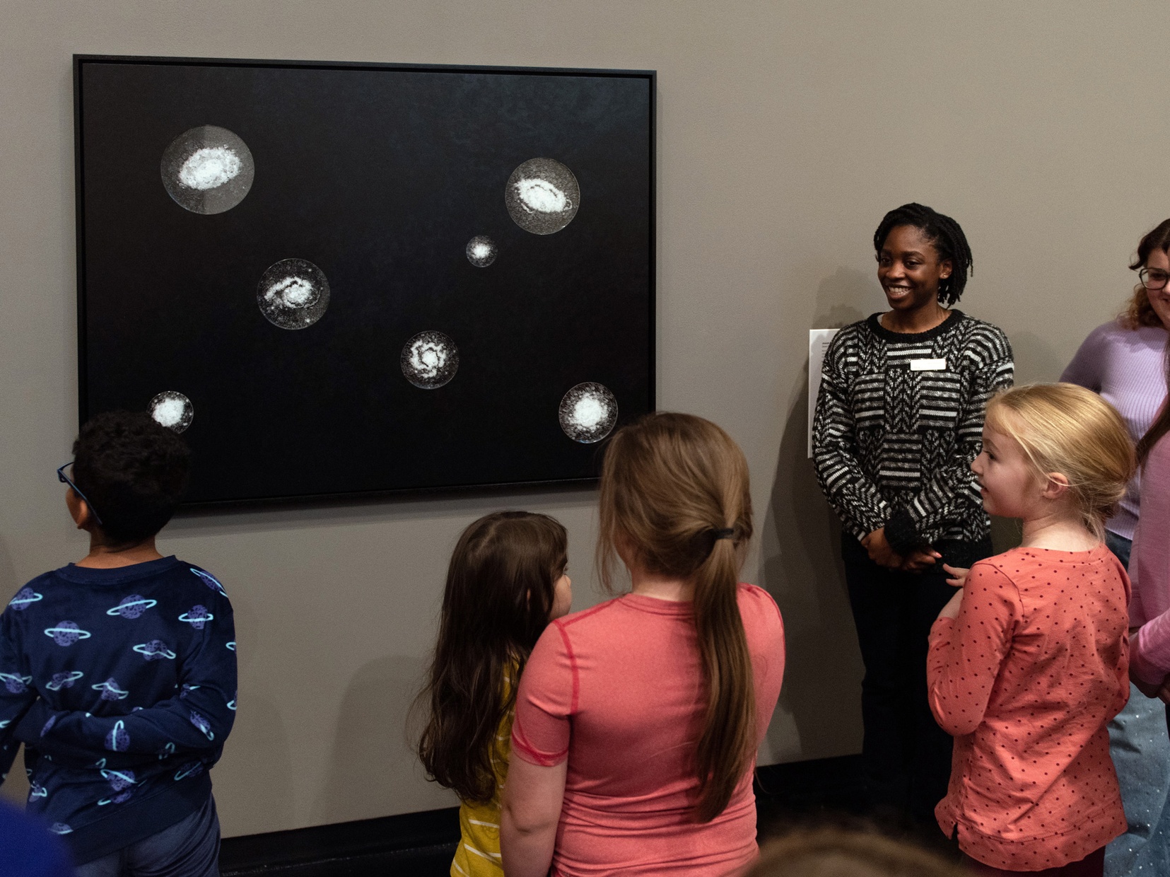 A Black woman stands next to an artwork hanging on the wall and looks out towards a group of children with their backs to us, observing the artwork.