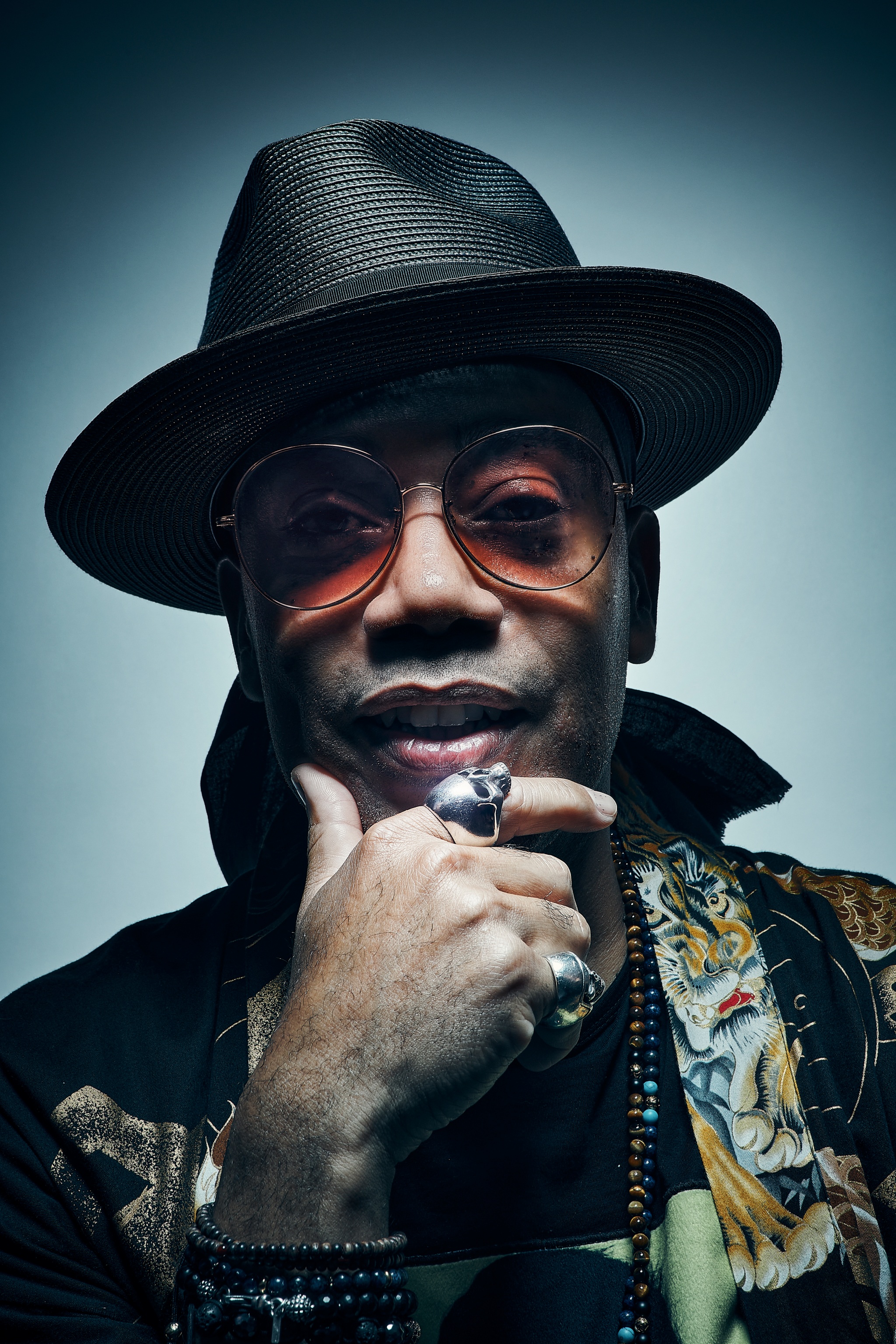 A Black man wearing orange sunglasses and a wide brimmed hat holds one hand to his chin. He wears two large rings on his pointer finger and pinky and looks directly at us from under the brim of his hat.