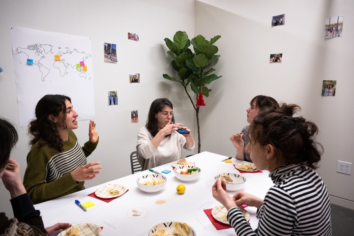 A group of people sit around a white table in a studio space and discuss something over small bowls of hummus.