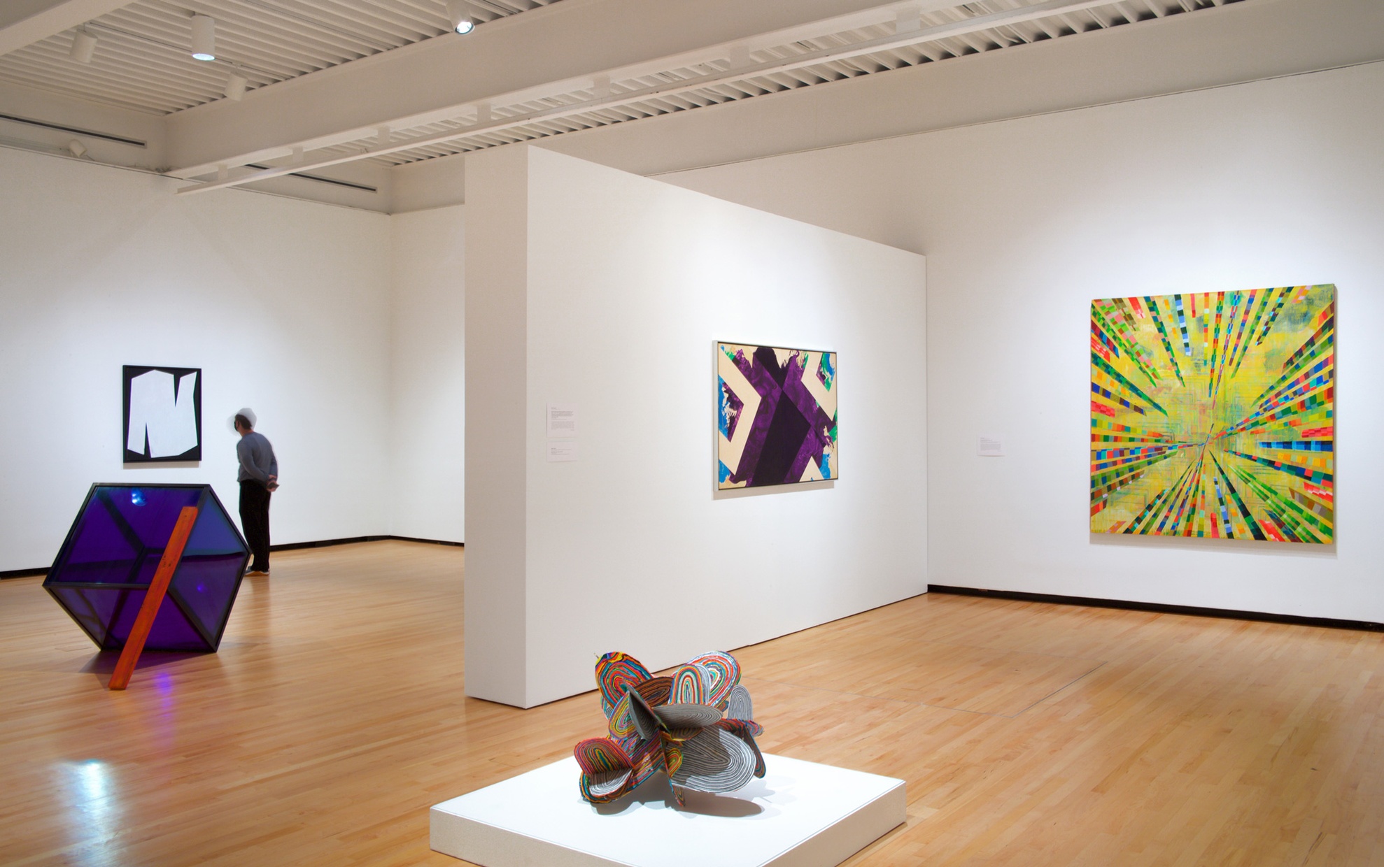 Colorful artwork hangs on white walls in a large room with sculptures sitting on both a pedestal and directly on the wood floors.