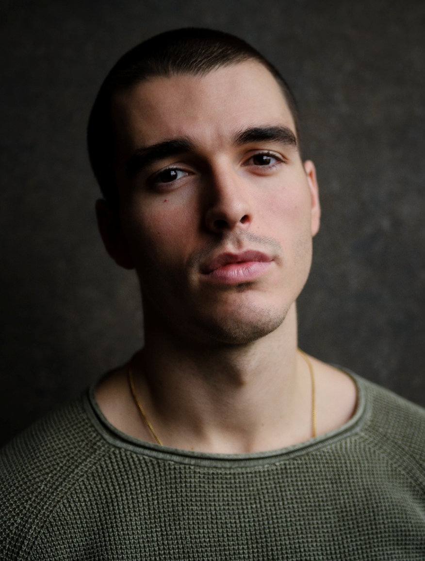 A headshot of actor Corey Mylchreest who angles his head back slightly while looking directly at us. He is white with short buzzed dark brown hair. He wears a khaki green crewneck shirt. 
