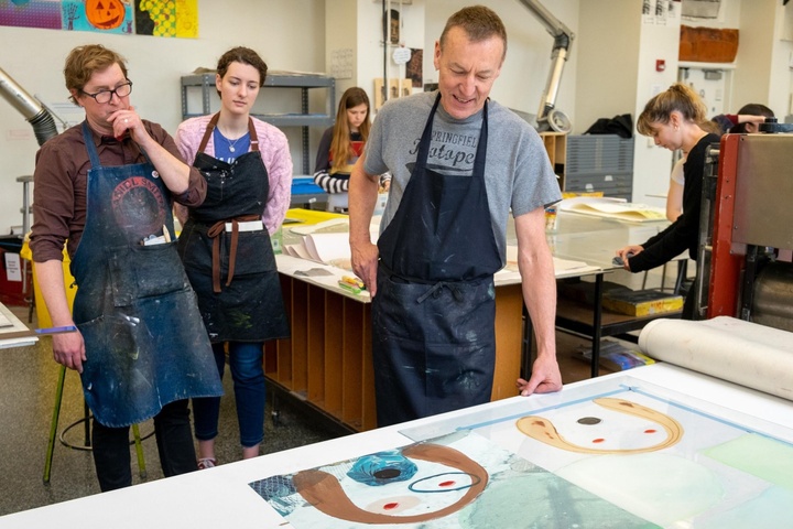 Three people review prints sitting on a printing press table. One person is smiling, another is touching their chin with their hand contemplatively. Behind them, other people are busy working around a large table. There are colorful prints on the walls, flat files, and ducts for ventilation in the background.