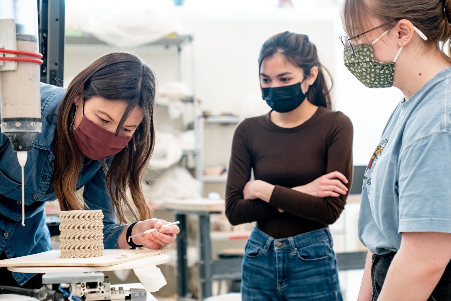 Three people wearing face coverings gather around a machine being used to make a 3D printed ceramics piece, which has a circular shape.