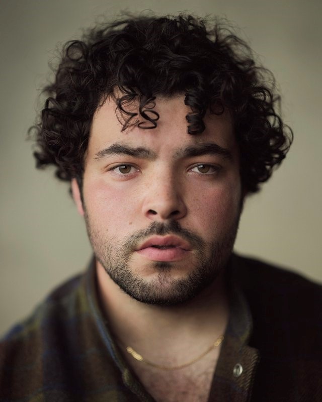 A headshot of actor Dylan Corbett-Bader, who looks directly at us with a slightly furrowed brow. He is white and has curly dark hair that falls down to the top of his cheeks. 