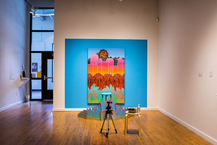 Small gallery space with one wall taken up by a teal blue painted photo backdrop depicting tropical foliage, lava, a blank neon cityscape, and fiery explosions in the distance. A painted stool is set in front with a little painted screen of foliage to hide the sitter's feet. An empty camera tripod is set up facing the display.