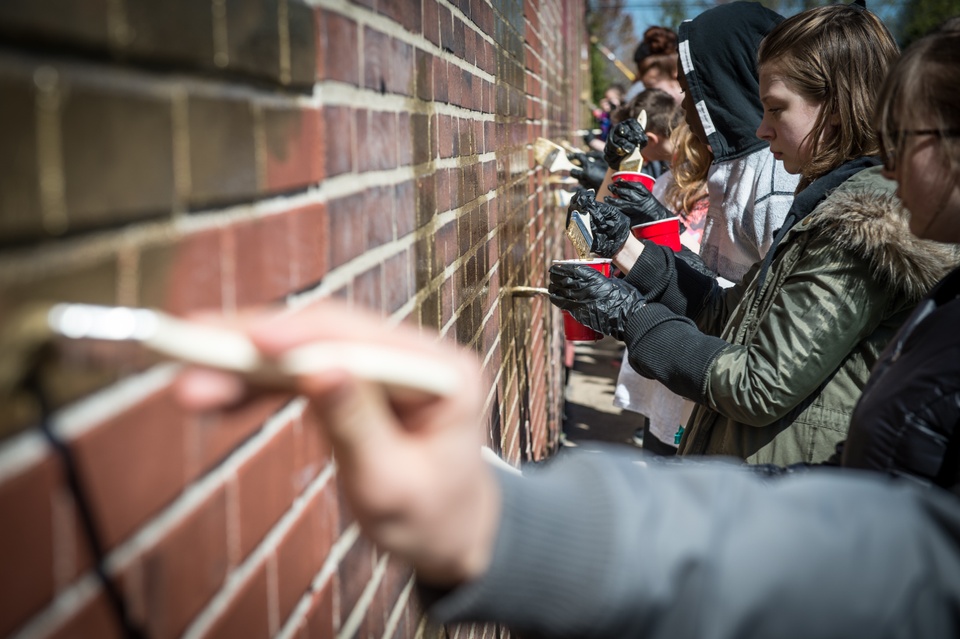 A group of people paint a brick wall gold with paintbrushes. 