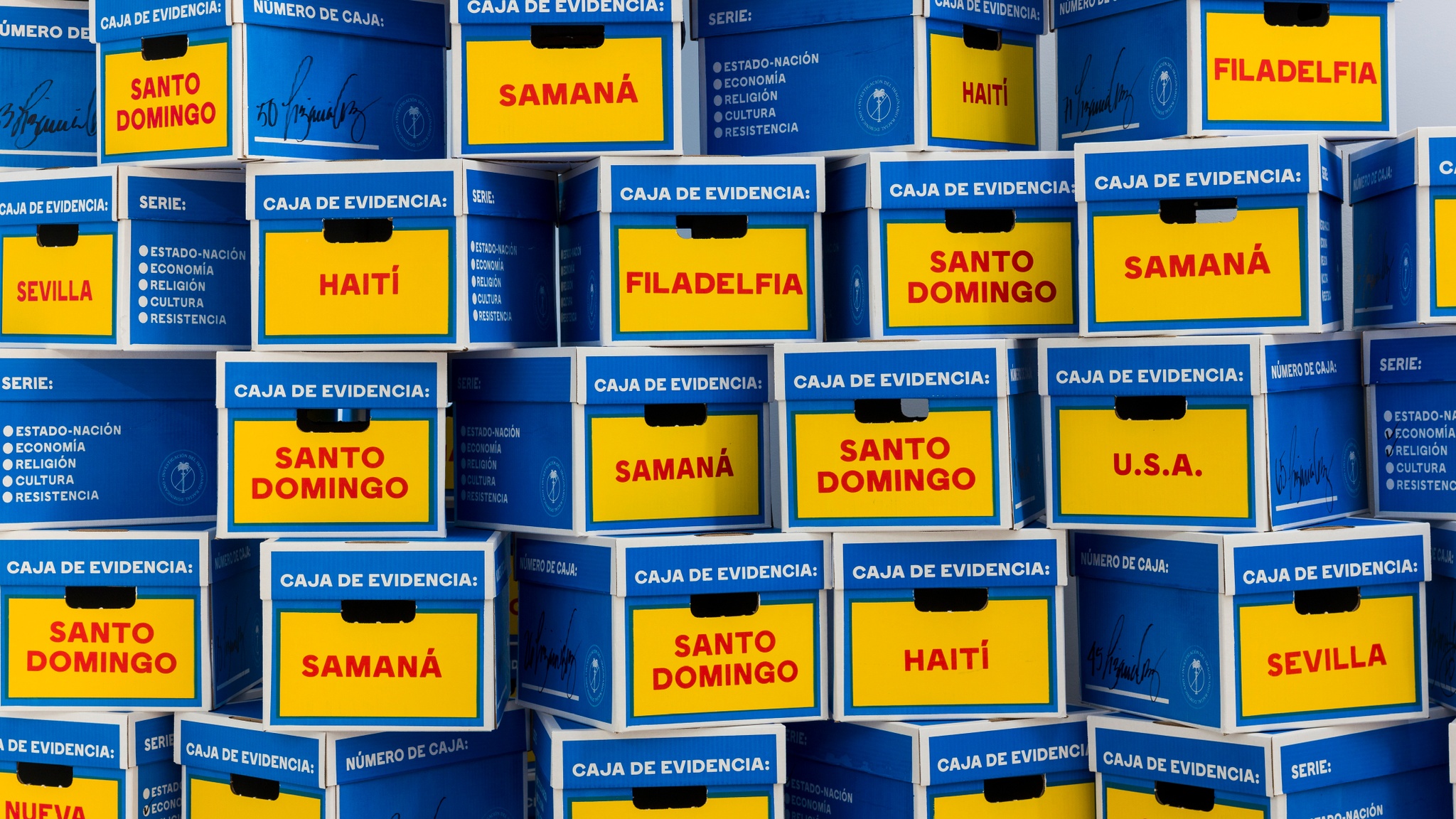 A stack of blue and yellow, cardboard evidence boxes, labeled in Spanish “Caja de evidencia.” The boxes are arranged in staggered columns and rows to create a wall of boxes. Each box is labeled with the name of a location in Spanish in red text, including Haití, Samana, Filadelfia, Santo Domingo, Nueva York, and Sevilla.