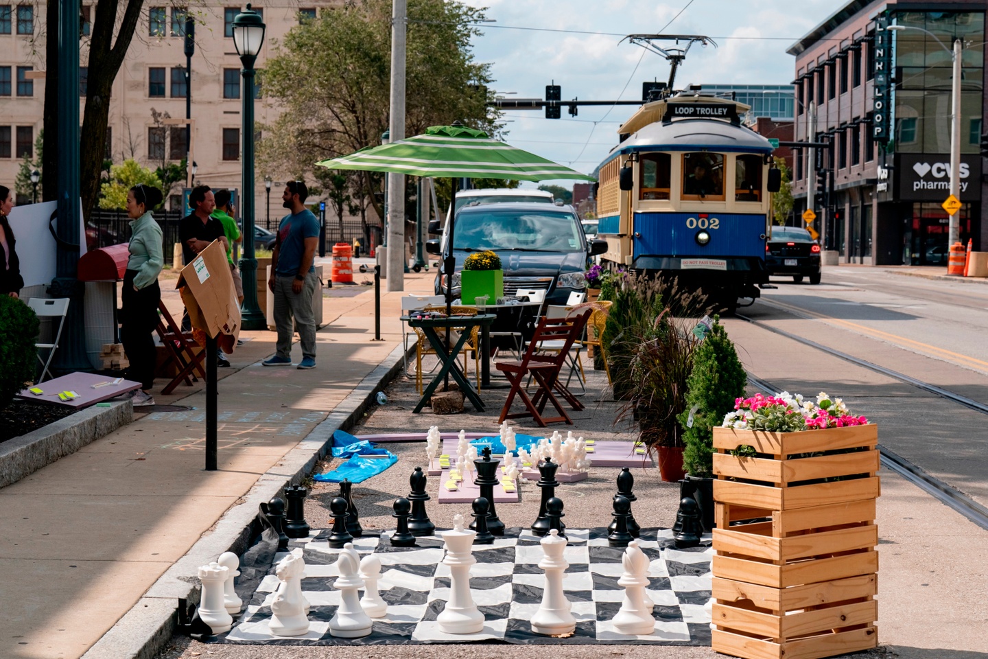 A large chess board, patio bistro set, and planters sit in a street in a parking spot as a trolley drives by.