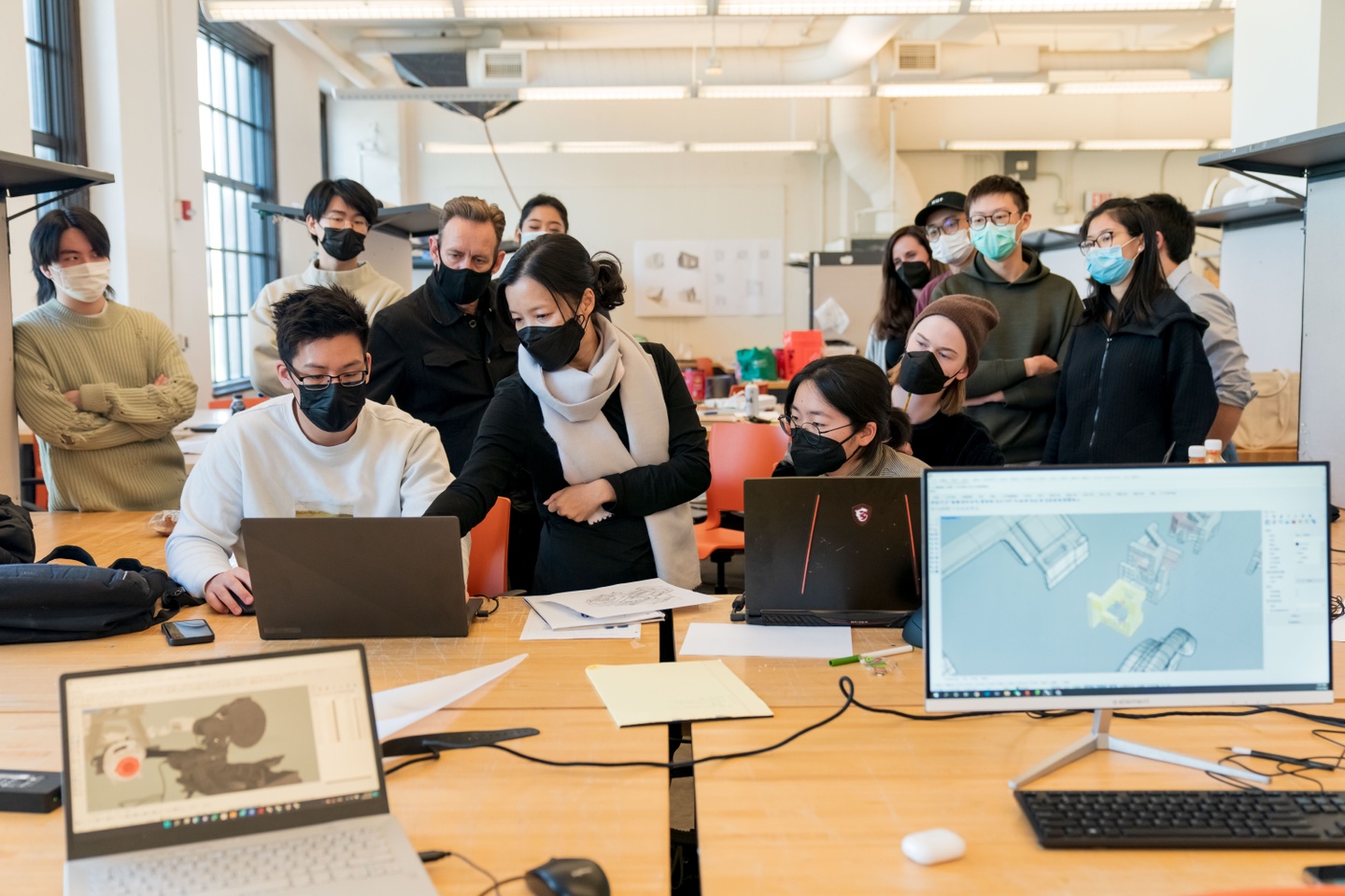 A group of students and faculty is gathered in an open studio space, looking at something on a student's computer screen. In the foreground of the photos are two computer screens, each with an architectural rendering.